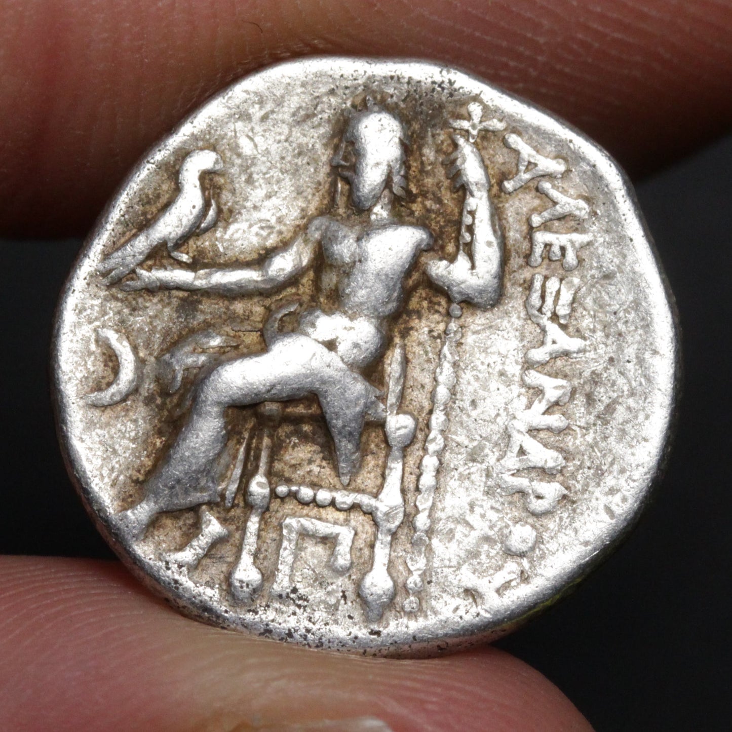 Ancient coin with a figure and text, held between fingers against a dark background