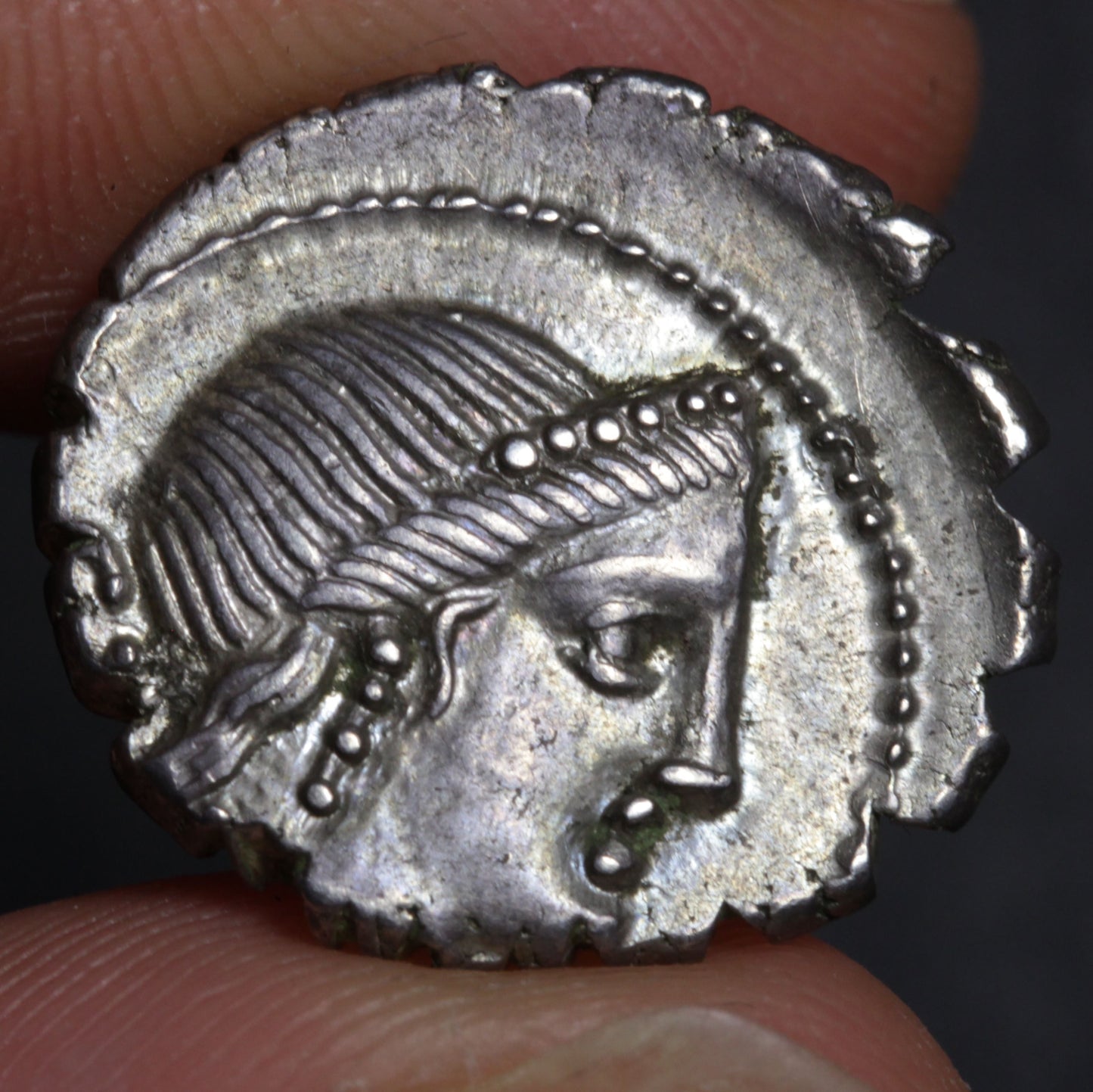 Ancient silver coin with a face design held between fingers against a dark background