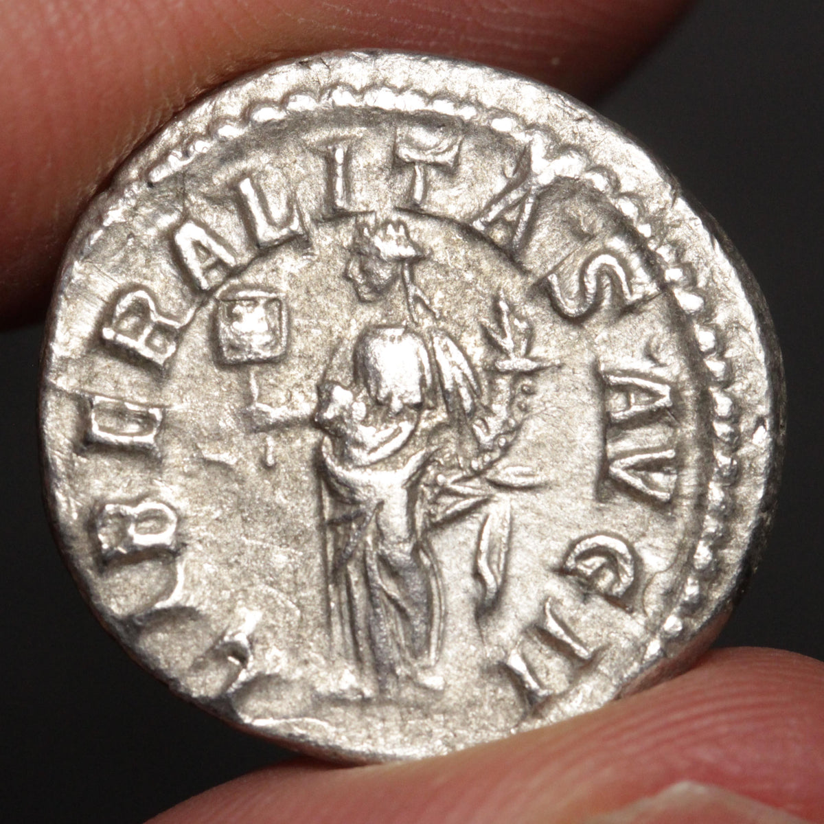 ancient silver coin with goddess standing and inscriptions, being held between two fingers against a dark background
