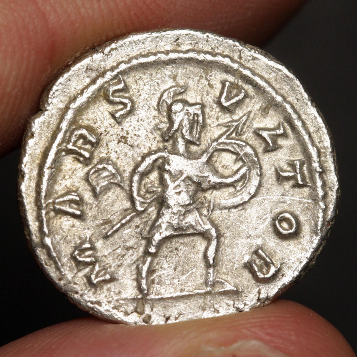 A close-up of an ancient silver coin held between fingers, showing a relief of a standing warrior holding a spear and shield, with Greek letters around the border.