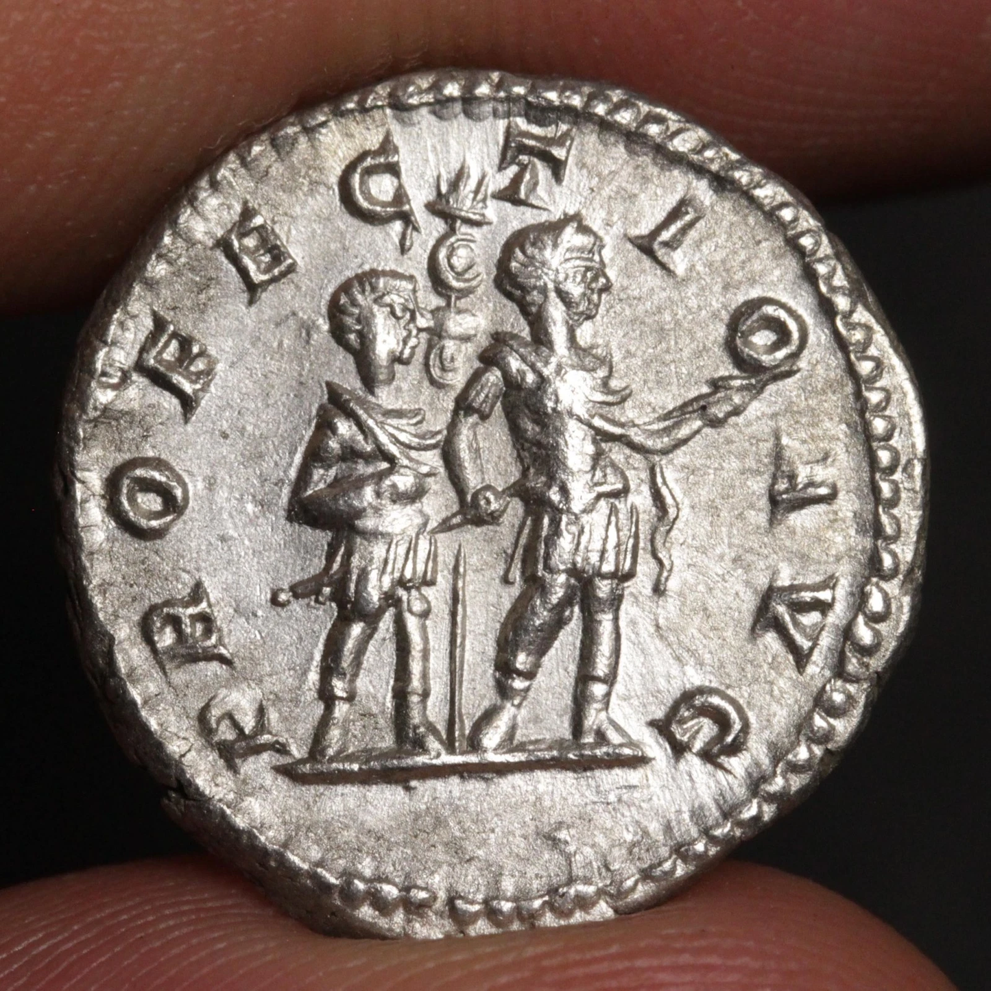 An ancient silver coin held between fingers, featuring two standing figures and raised Roman numerals around the edge.