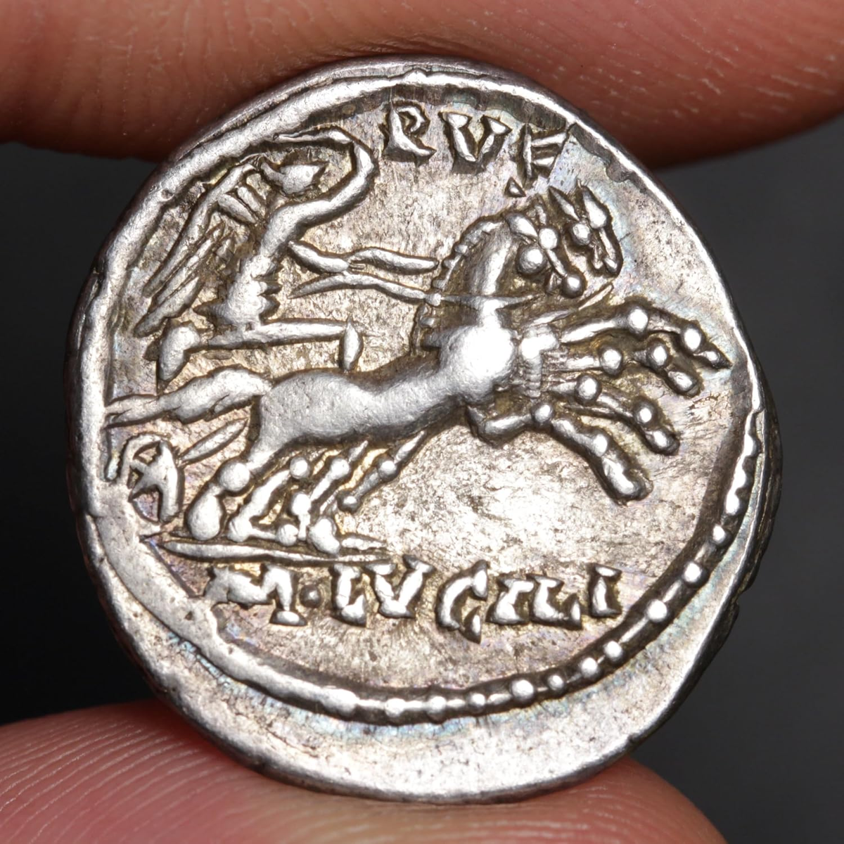 An ancient silver-toned coin featuring a rearing horse and rider design, held between two fingers against a dark background.
