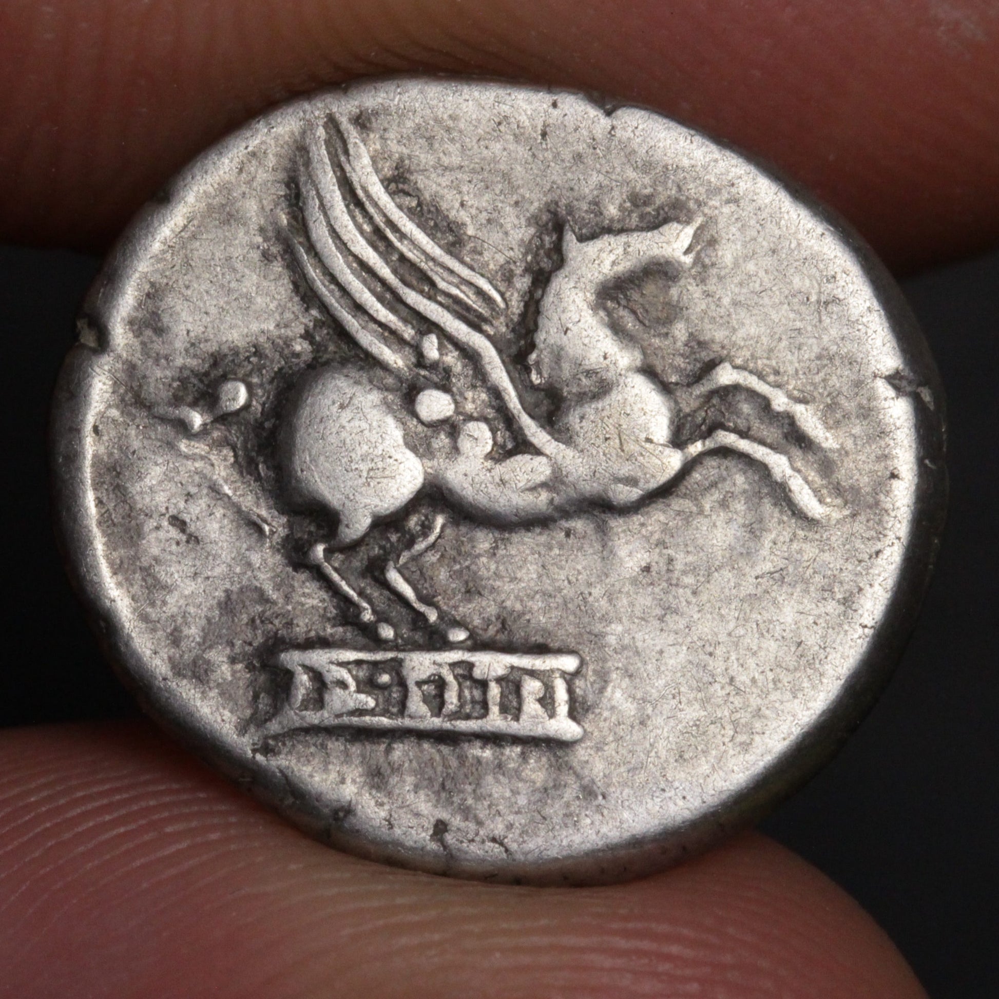 Ancient coin with a winged horse and text, held between fingers