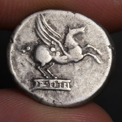 Ancient coin with a winged horse and text, held between fingers