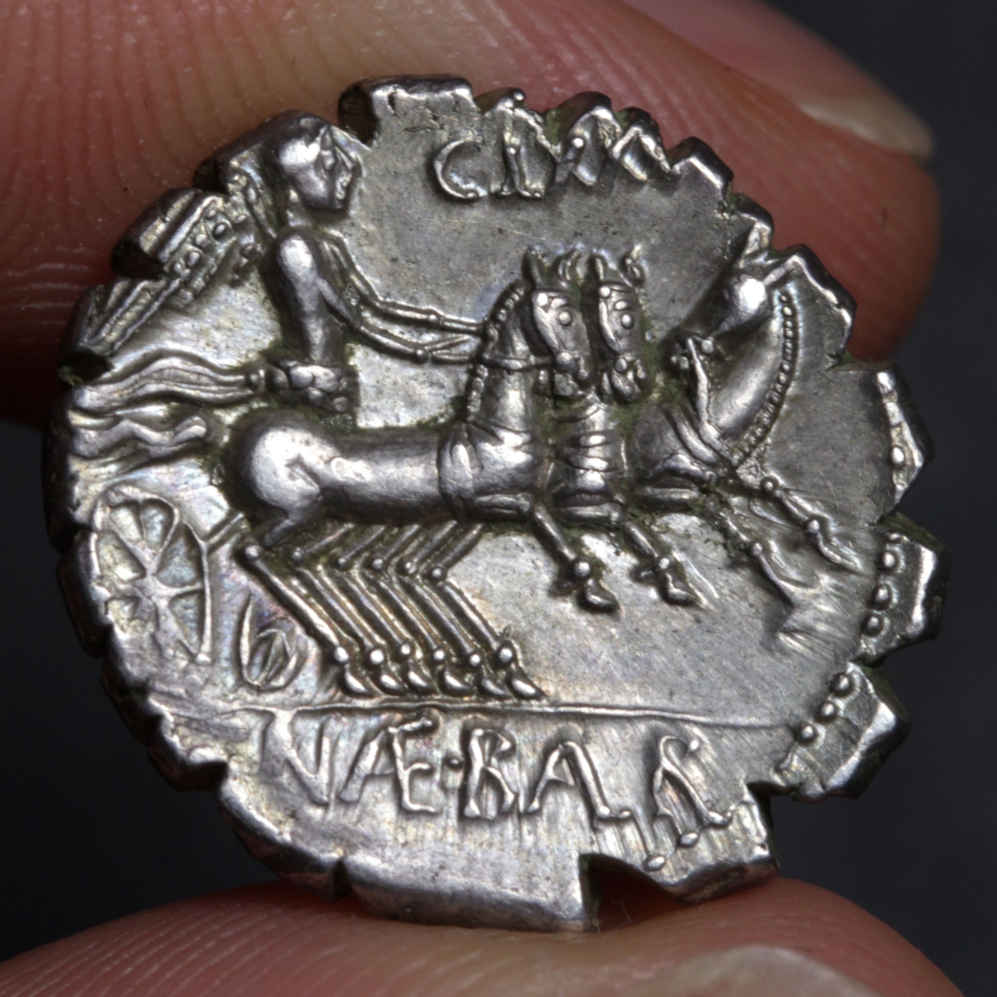 Silver coin with a chariot design held between fingers against a dark background