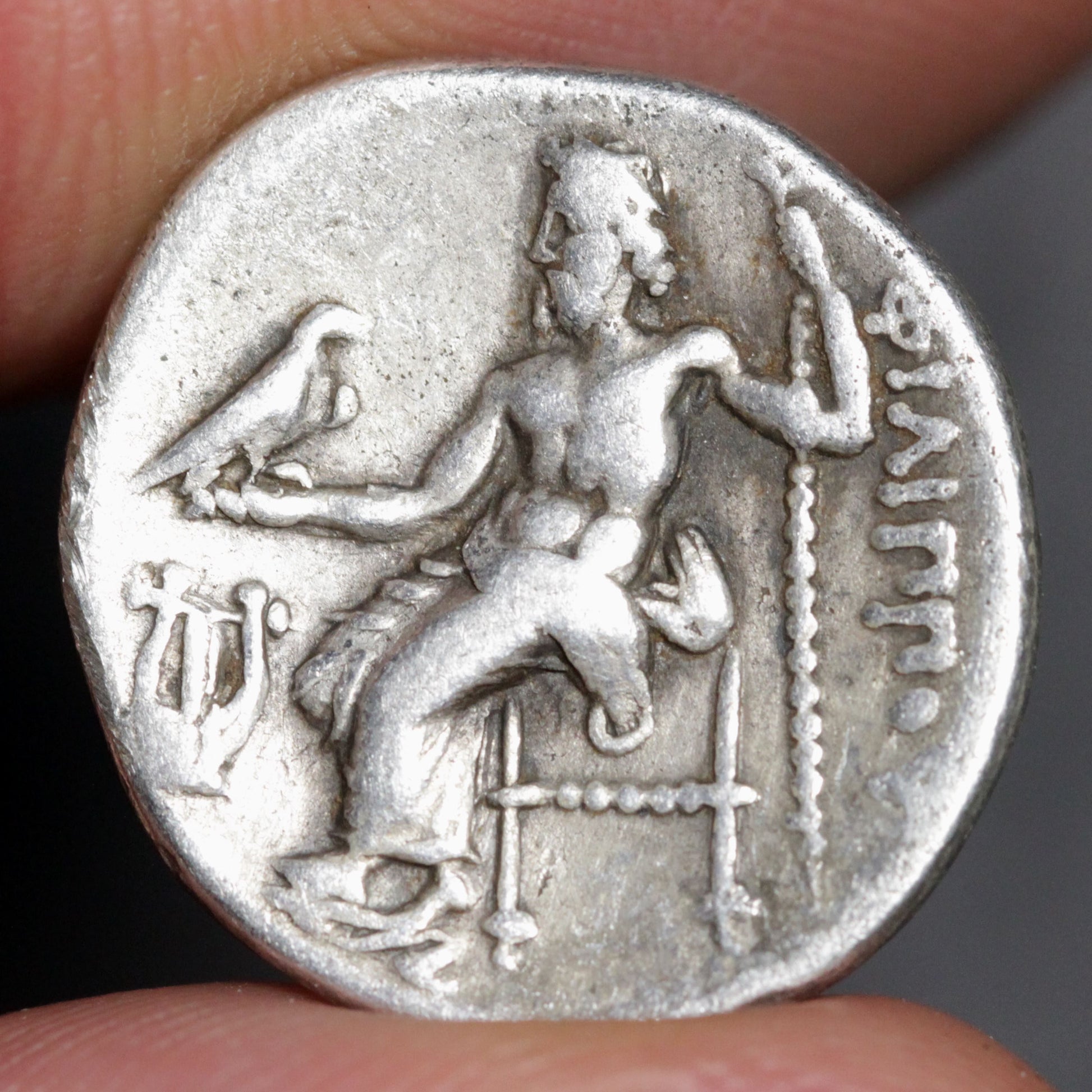 Ancient silver coin with a seated figure and inscriptions held between fingers