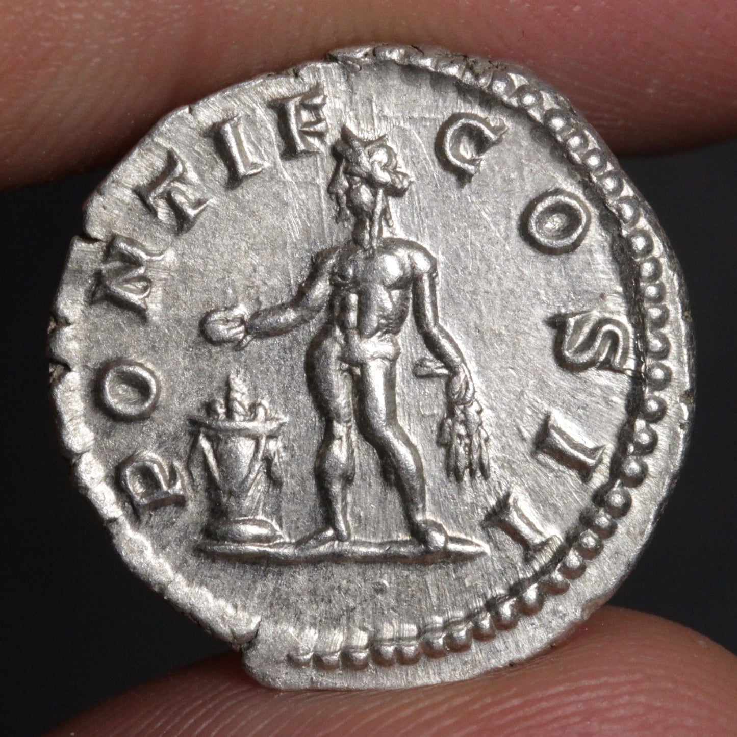 An ancient silver coin featuring a standing figure and the text 'PONTIF COS', held between fingers against a dark background.