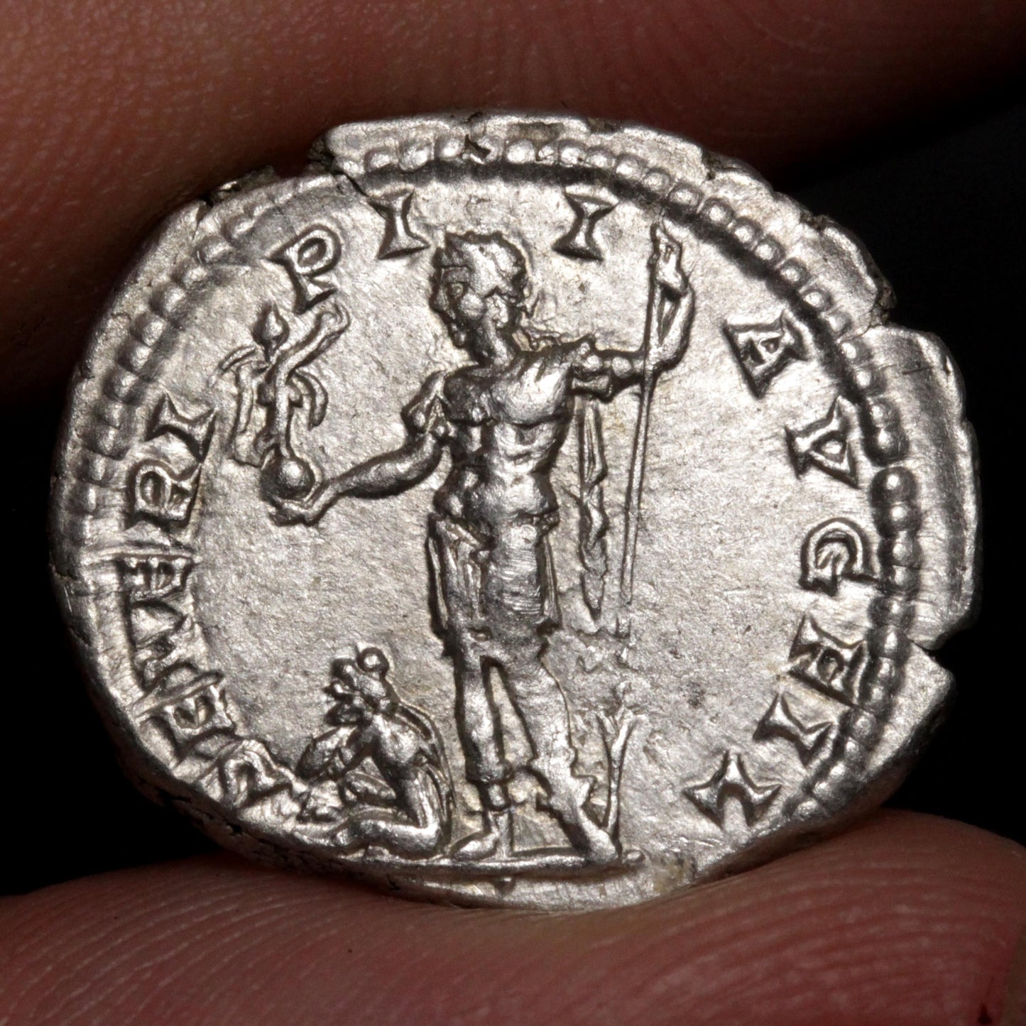 An ancient silver coin held between fingers, showing a detailed relief of a standing figure holding a staff and a Victory, with inscribed text around the edge.