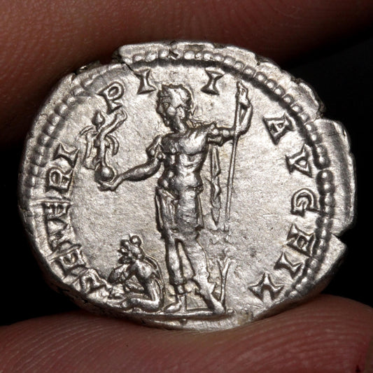 An ancient silver coin held between fingers, showing a detailed relief of a standing figure holding a staff and a Victory, with inscribed text around the edge.