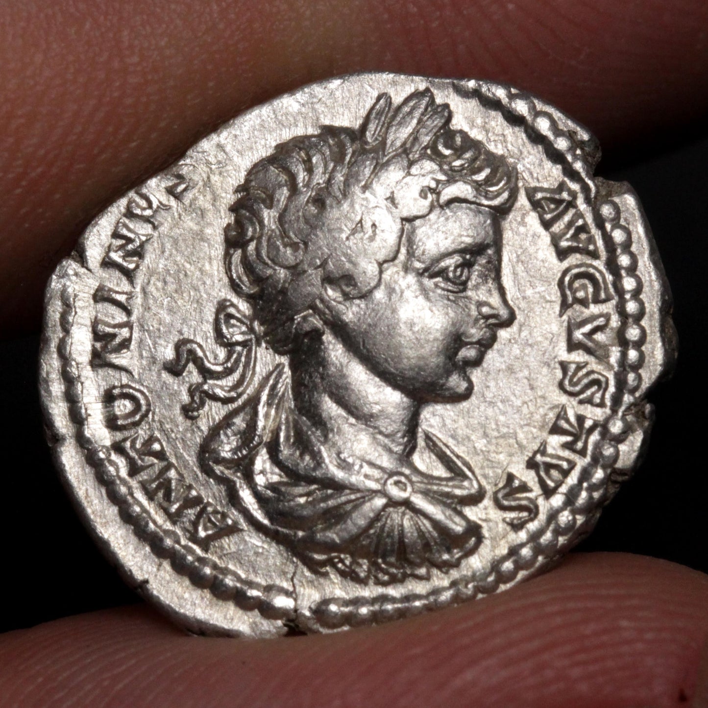 An ancient silver coin featuring a profile portrait of a Roman emperor, held between human fingers against a dark background.