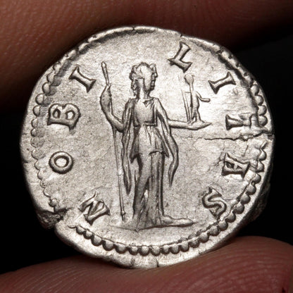 An ancient silver coin featuring a standing figure with outstretched arms, held between fingers against a dark background.