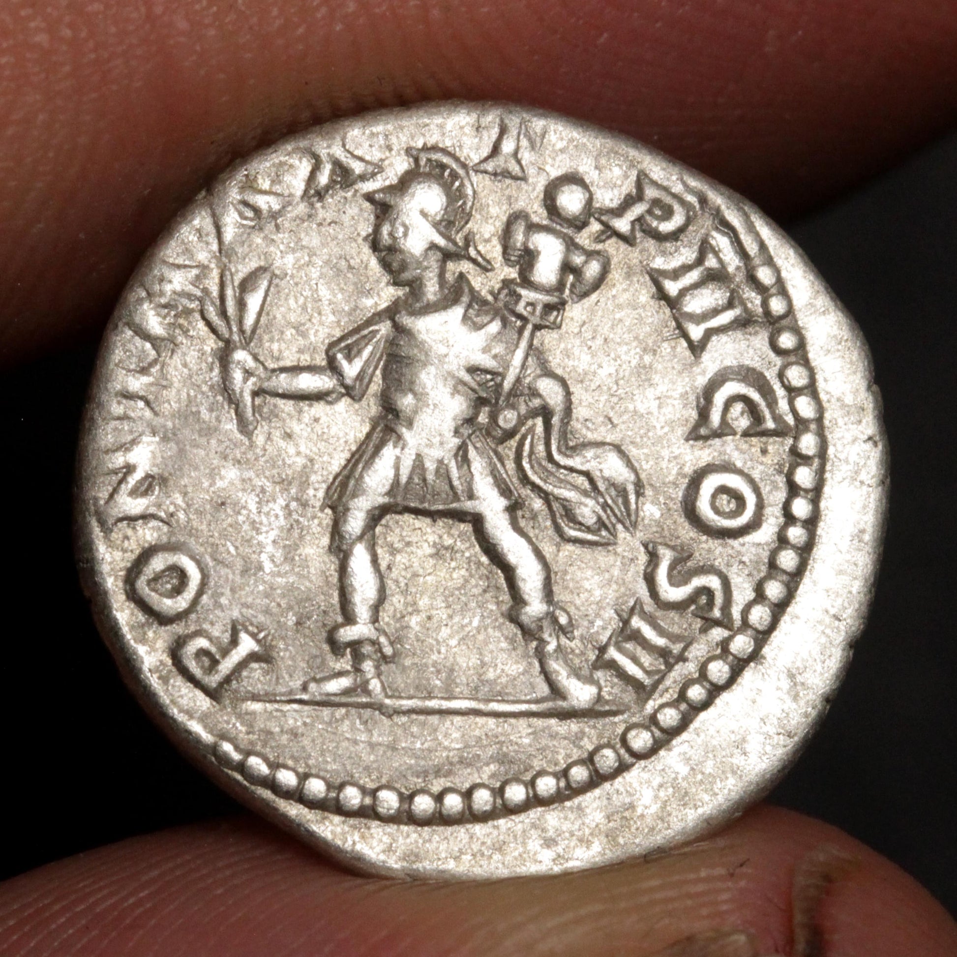 An ancient silver coin featuring a standing figure, held between fingers against a dark background.
