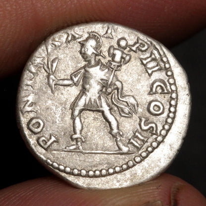 An ancient silver coin featuring a standing figure, held between fingers against a dark background.