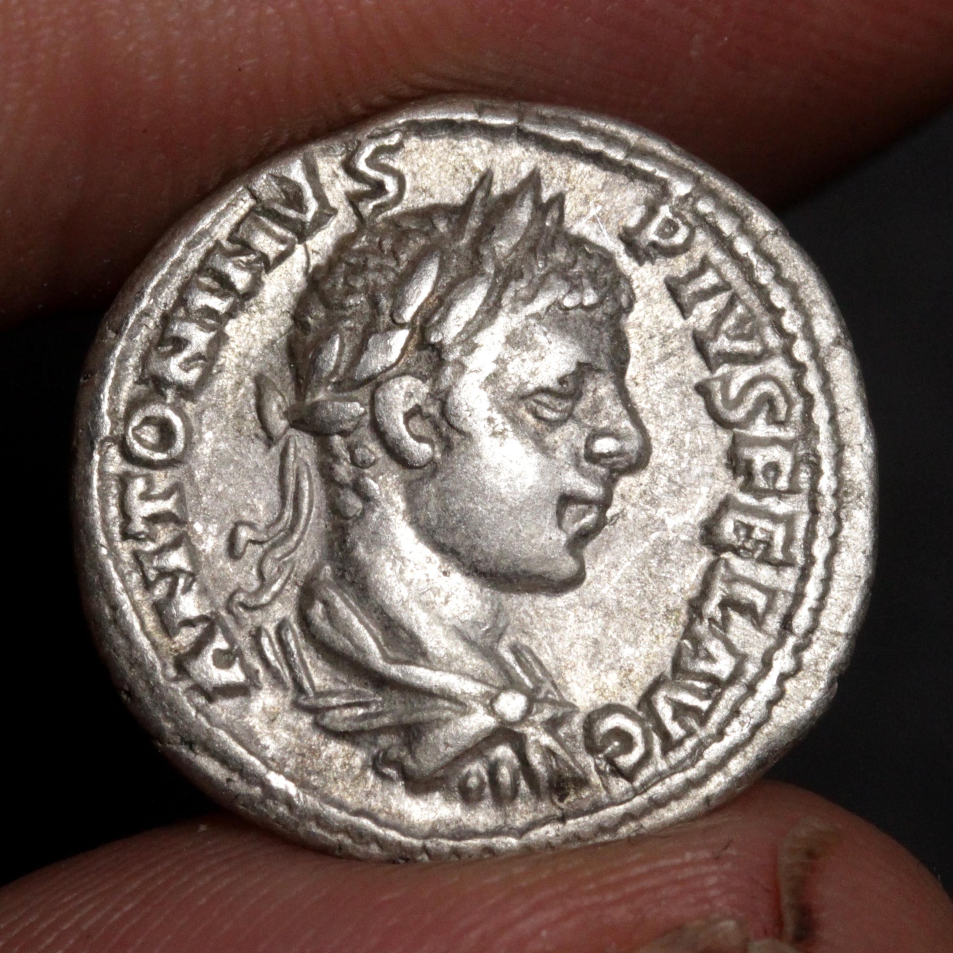 An ancient silver coin featuring a profile portrait of a Roman emperor, held between two fingers to show its scale.
