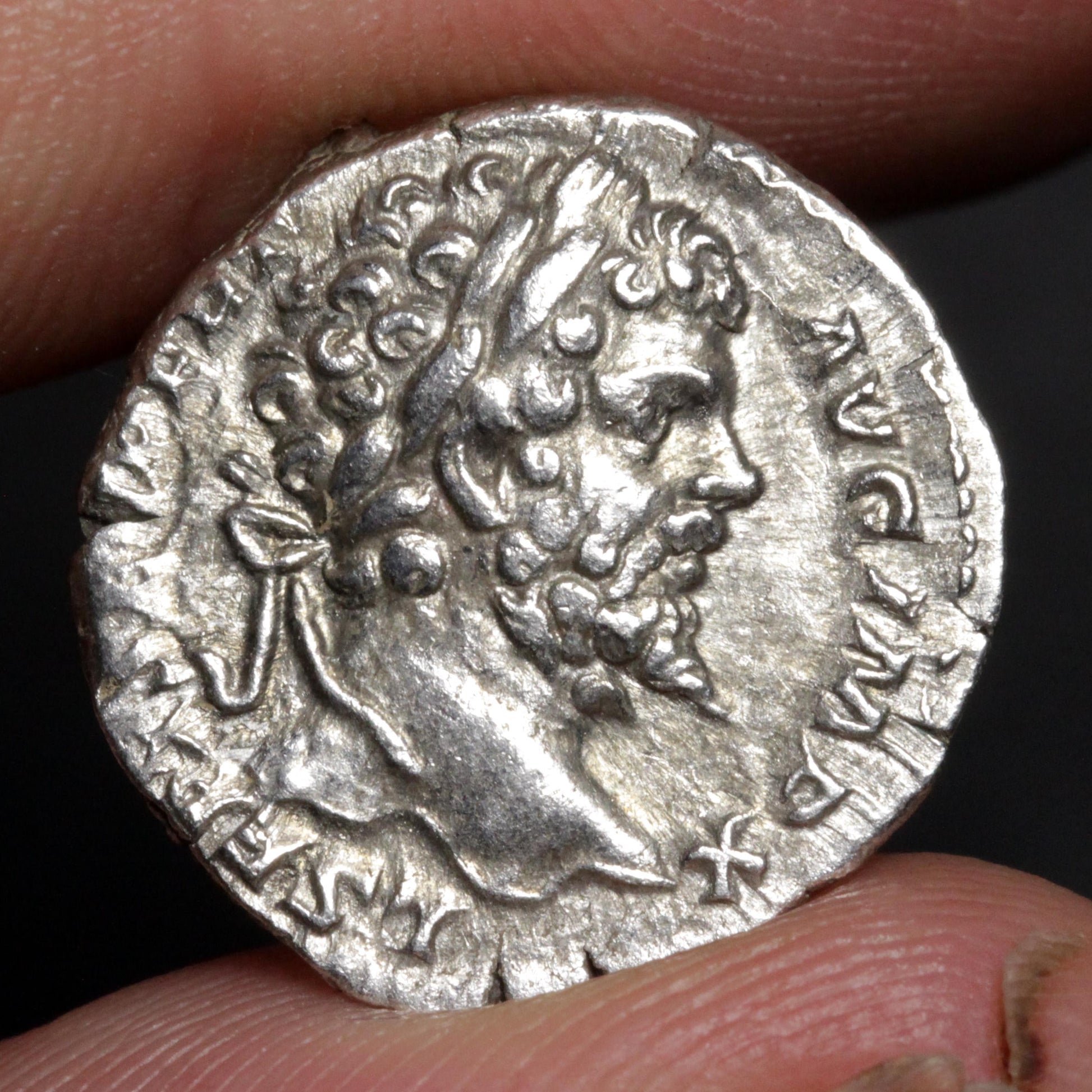 An ancient silver coin featuring a profile portrait of a bearded man, held between two fingers against a dark background.