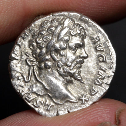 An ancient silver coin featuring a profile portrait of a bearded man, held between two fingers against a dark background.