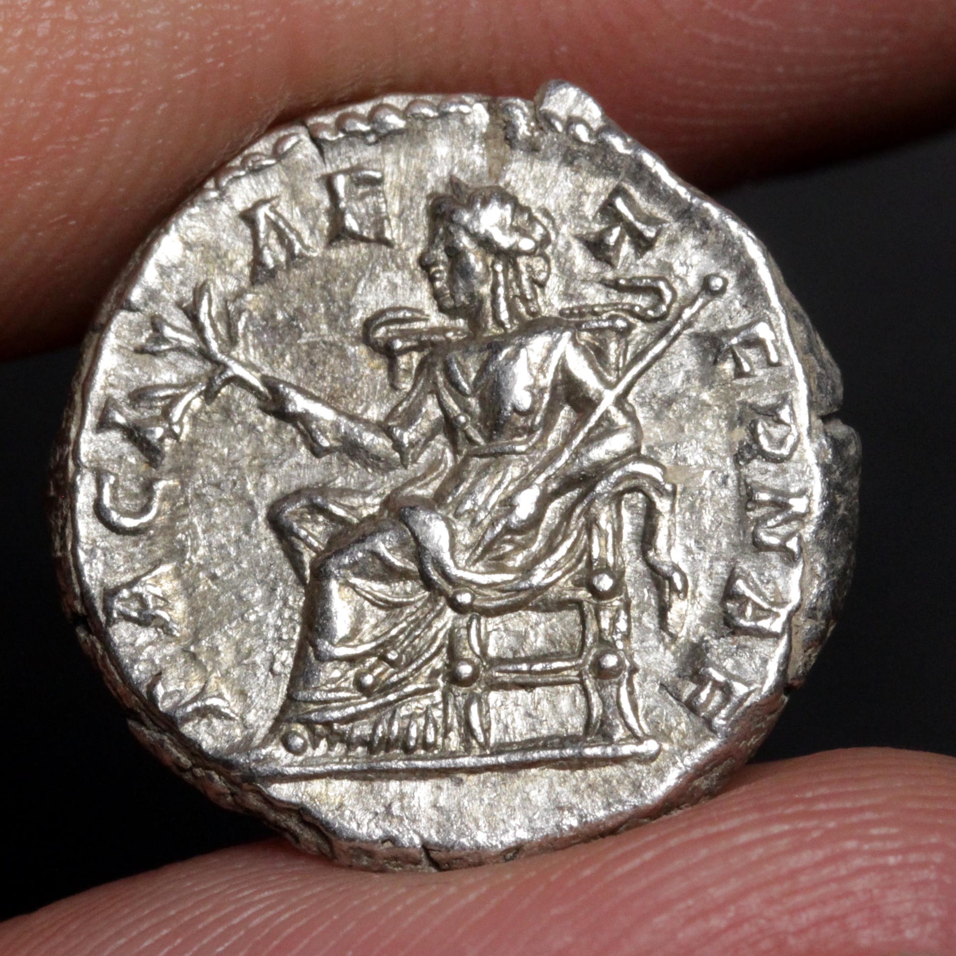 An ancient silver coin featuring a seated figure and Roman lettering, held between human fingers against a dark background.