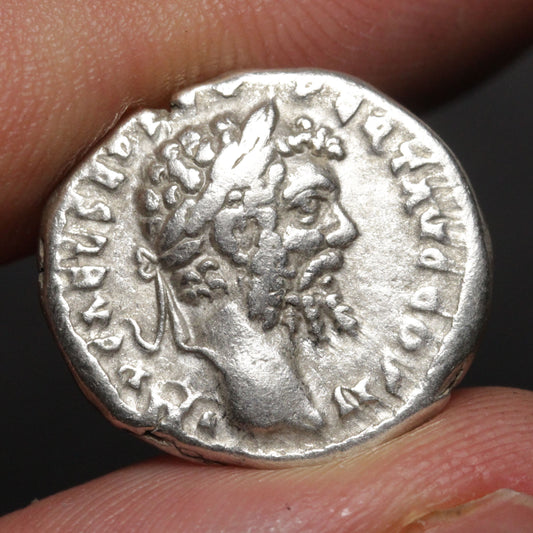 An ancient silver coin featuring a profile portrait of a bearded man wearing a laurel wreath, held between two fingers against a dark background.