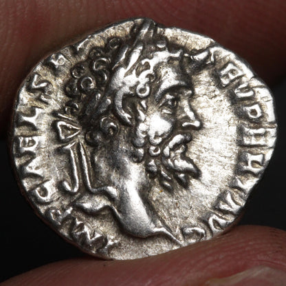 An ancient silver coin featuring a profile portrait of a bearded man, held between fingers against a dark background.