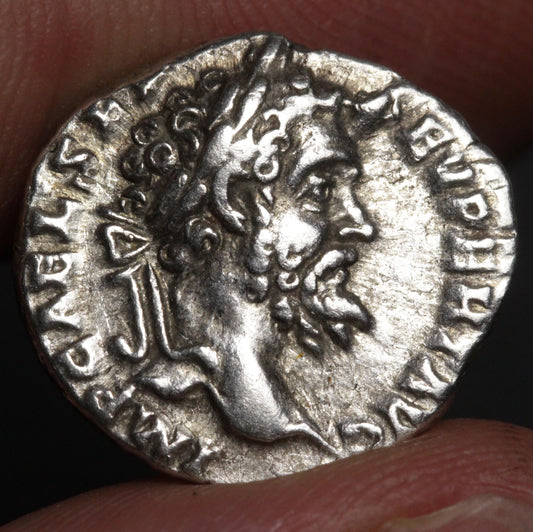 An ancient silver coin featuring a profile portrait of a bearded man, held between fingers against a dark background.