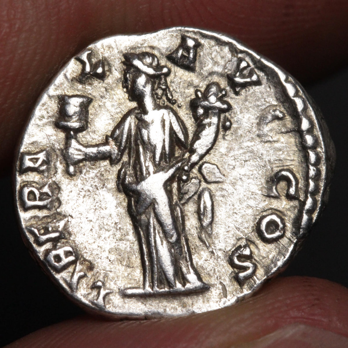 An ancient silver coin featuring a standing figure holding a cup and a branch, with the inscription around the edge, shown resting on fingers.