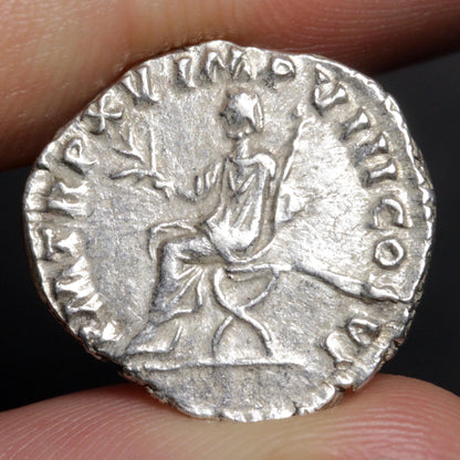 an ancient silver coin being held between two fingers showing a man seated and holding an olive-branch