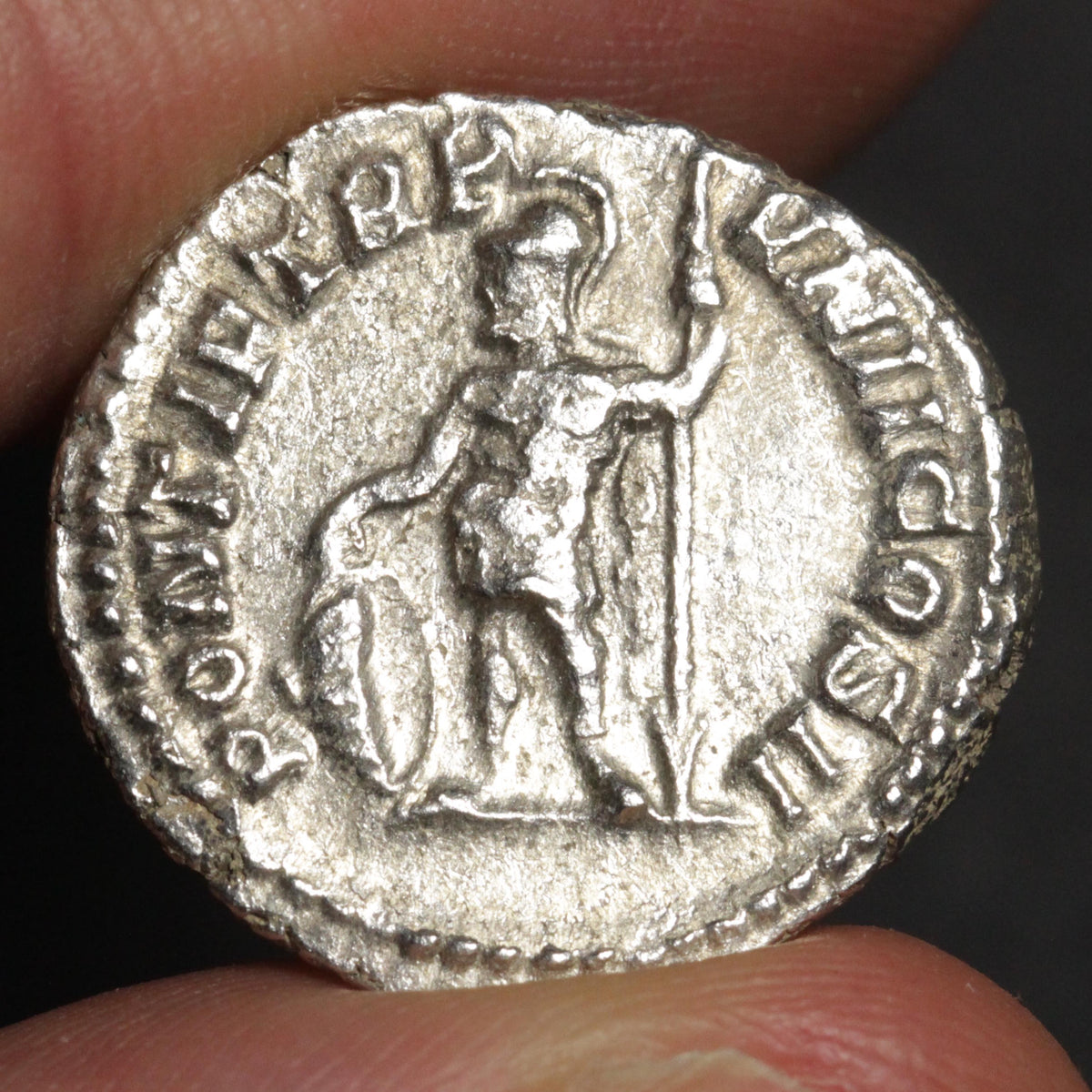 An ancient silver coin featuring a standing figure holding a shield and spear, with the inscription 'PORT IERBB' around the edge, held between fingers against a dark background.