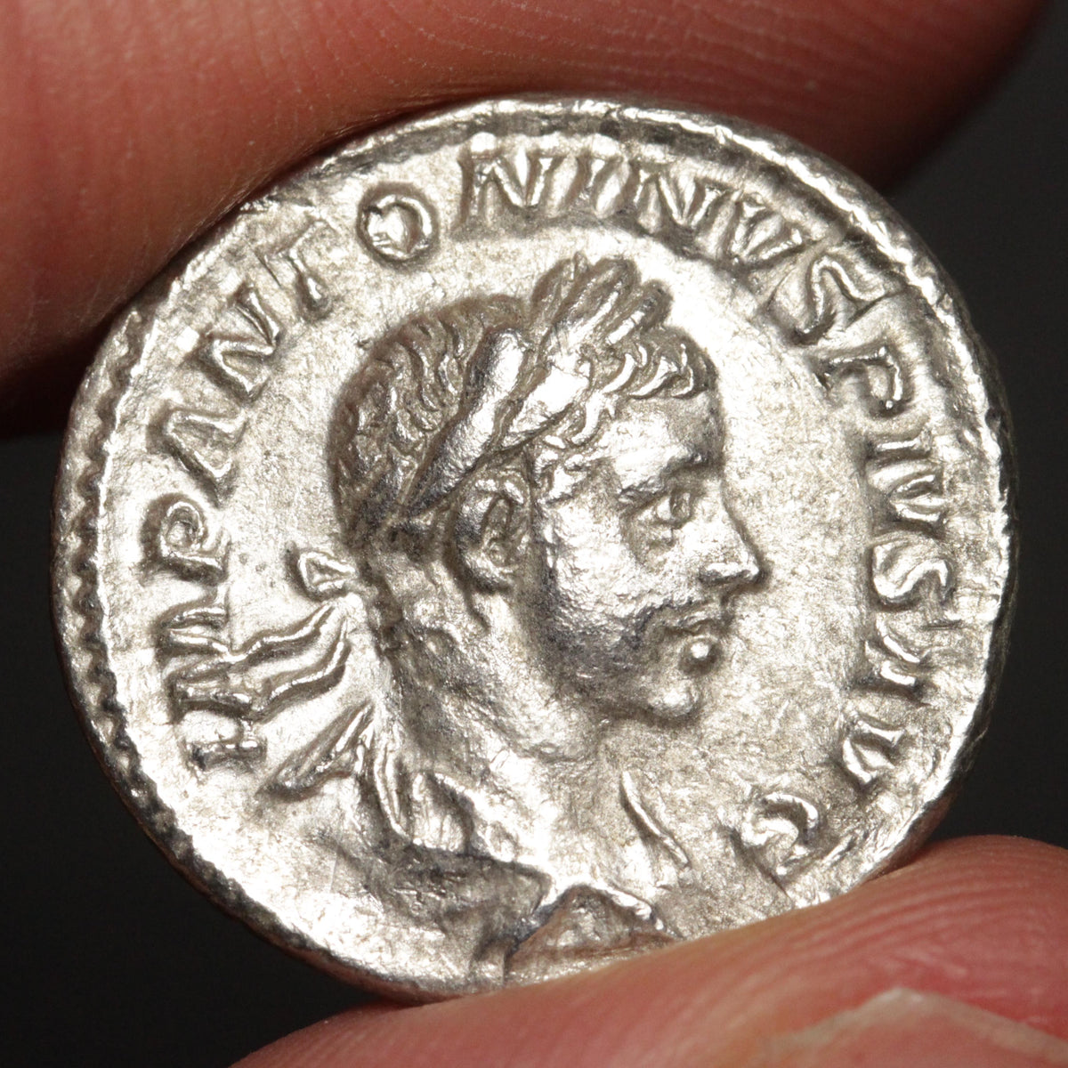 ancient silver coin with bust of young man, being held between two fingers against a dark background