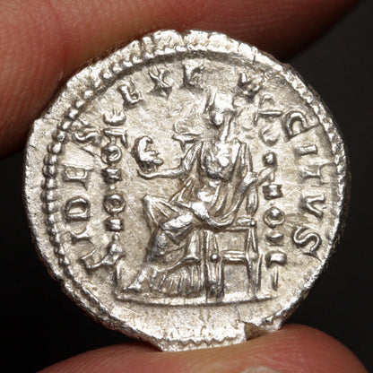 An ancient silver coin featuring a seated figure and raised inscription, held between fingers against a dark background.