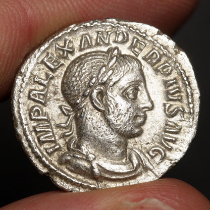 An ancient silver Roman coin featuring a profile portrait of a male figure, held between two fingers against a dark background.
