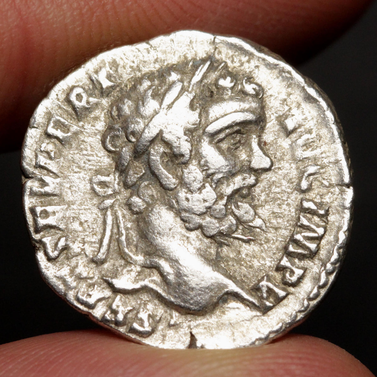 An ancient silver coin featuring a profile portrait of a bearded man, held between two fingers against a dark background.