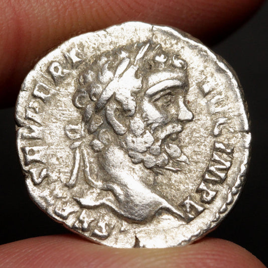 An ancient silver coin featuring a profile portrait of a bearded man, held between two fingers against a dark background.