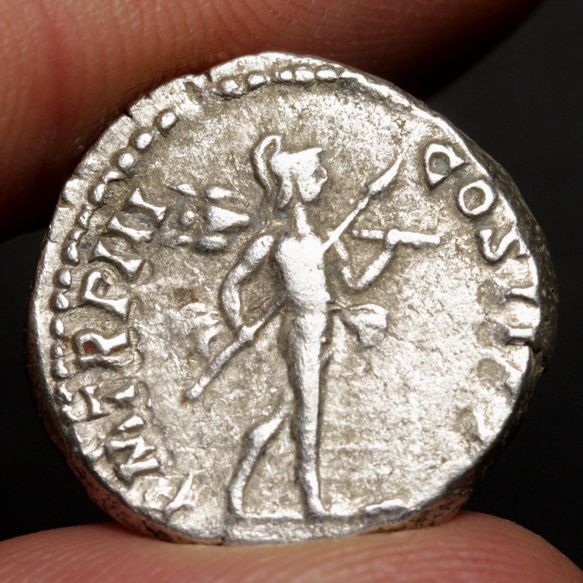 An ancient-style silver coin held between fingers, featuring a standing figure and raised inscription around the edge, photographed close-up against a dark background.