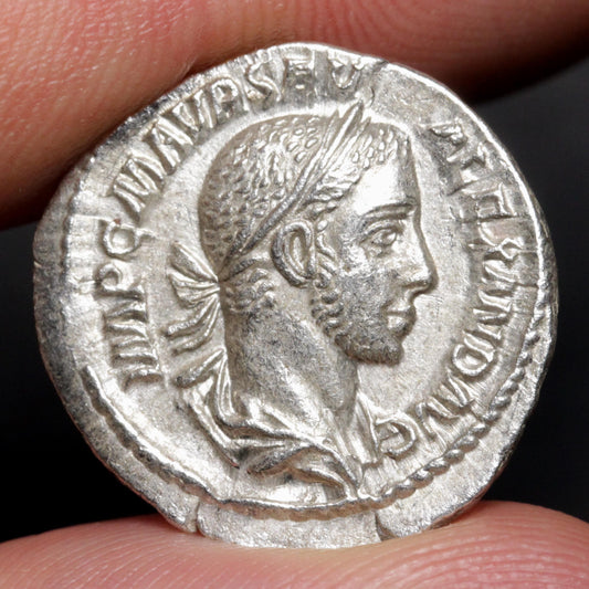 An ancient silver coin featuring a profile portrait of a Roman emperor, held between two fingers against a dark background.