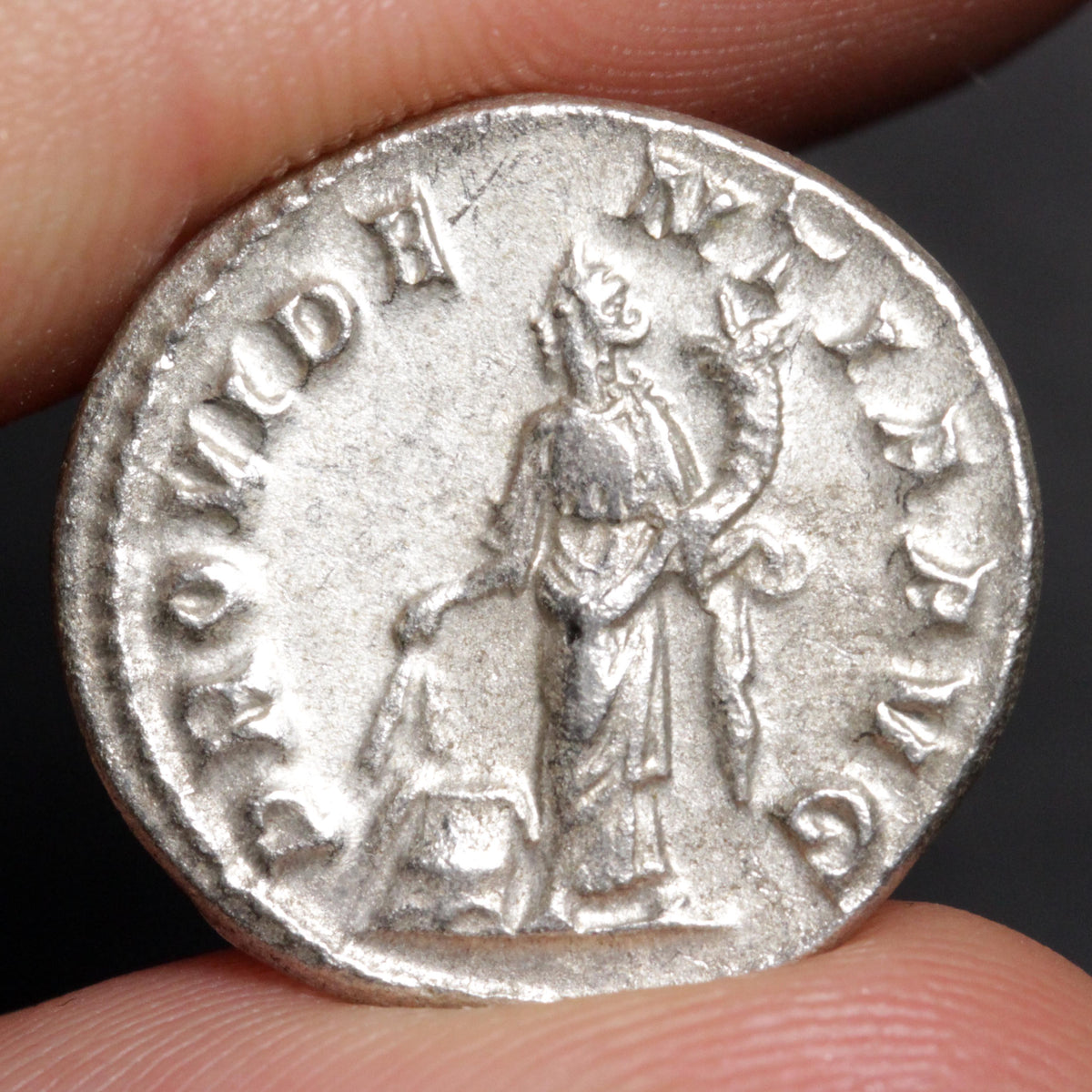 An ancient silver Roman coin featuring a standing figure and raised inscription, held between two fingers against a dark background.