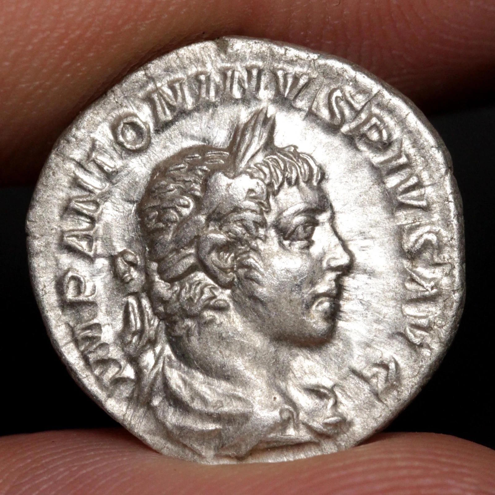 An ancient silver Roman coin featuring a profile portrait of a Roman emperor, held between two fingers to show scale against a dark background.