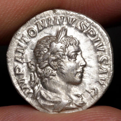 An ancient silver Roman coin featuring a profile portrait of a Roman emperor, held between two fingers to show scale against a dark background.