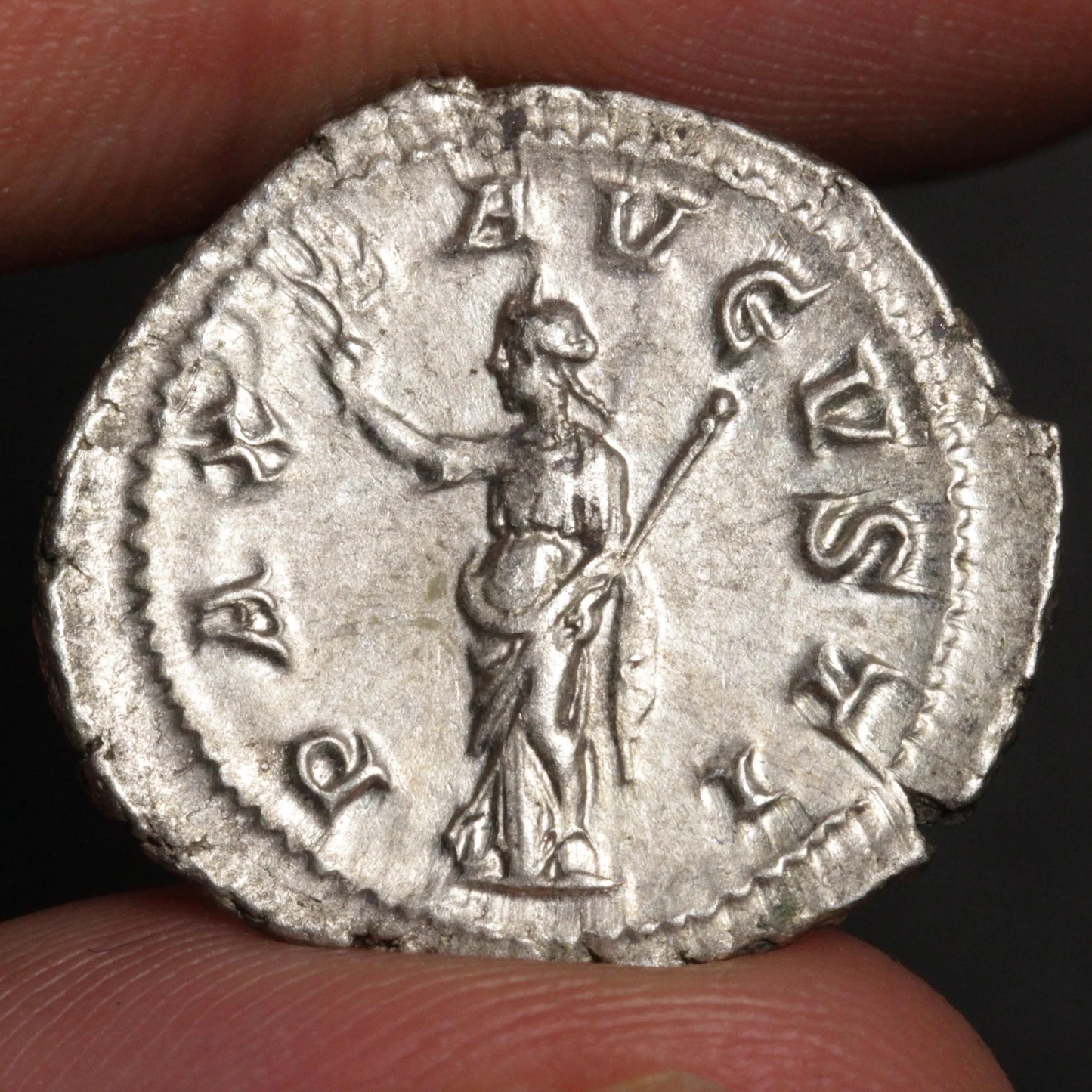 An ancient silver coin featuring a standing figure with a staff and torch, surrounded by raised letters, held between fingers against a dark background.