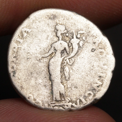 An ancient silver coin featuring a standing female figure, held between two human fingers against a dark background.