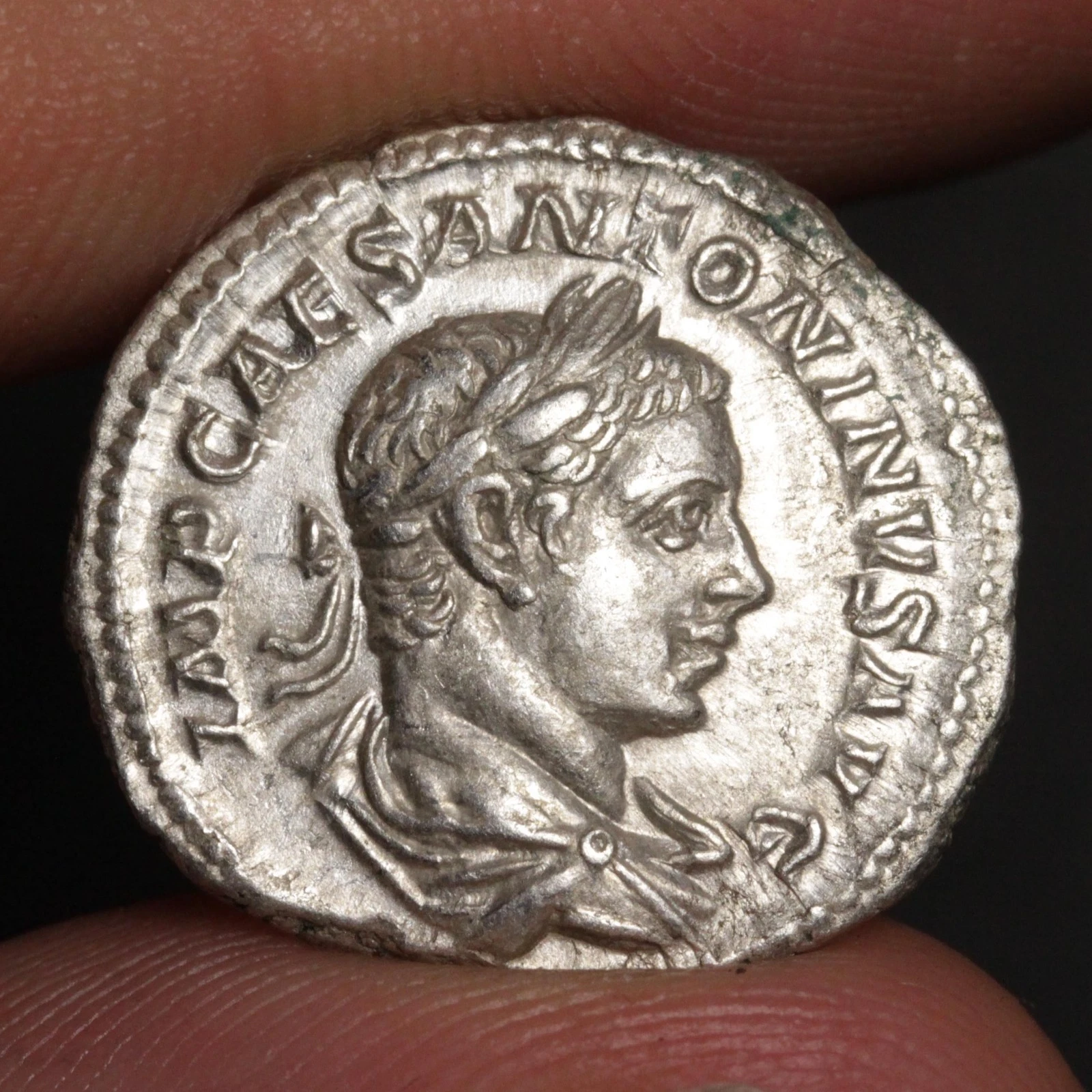 An ancient silver Roman coin featuring a profile portrait of a emperor, held between two fingers to show scale against a dark background.