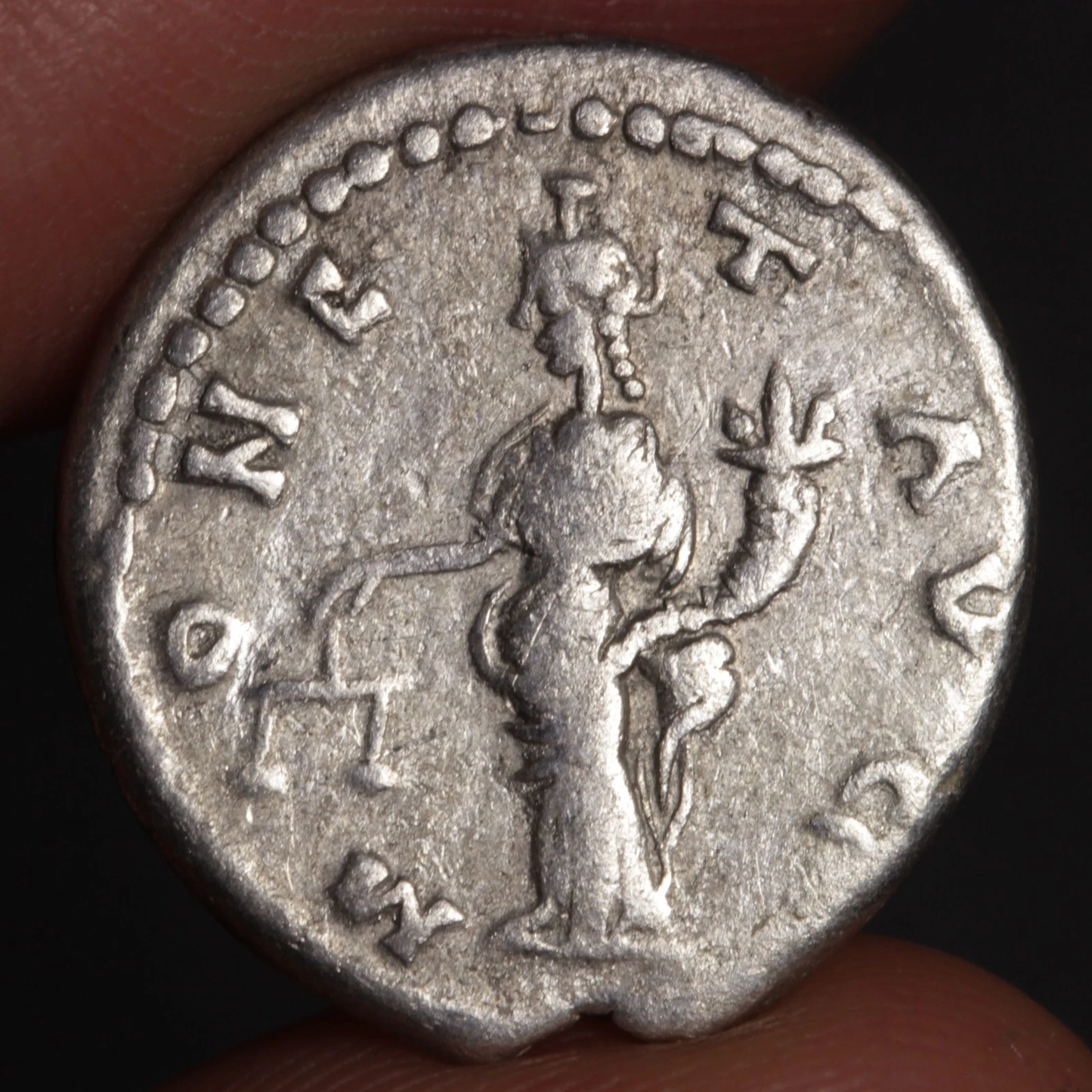 Ancient silver coin with a figure and inscriptions held between fingers against a dark background