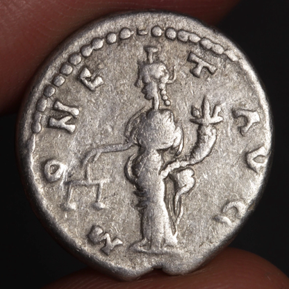 Ancient silver coin with a figure and inscriptions held between fingers against a dark background