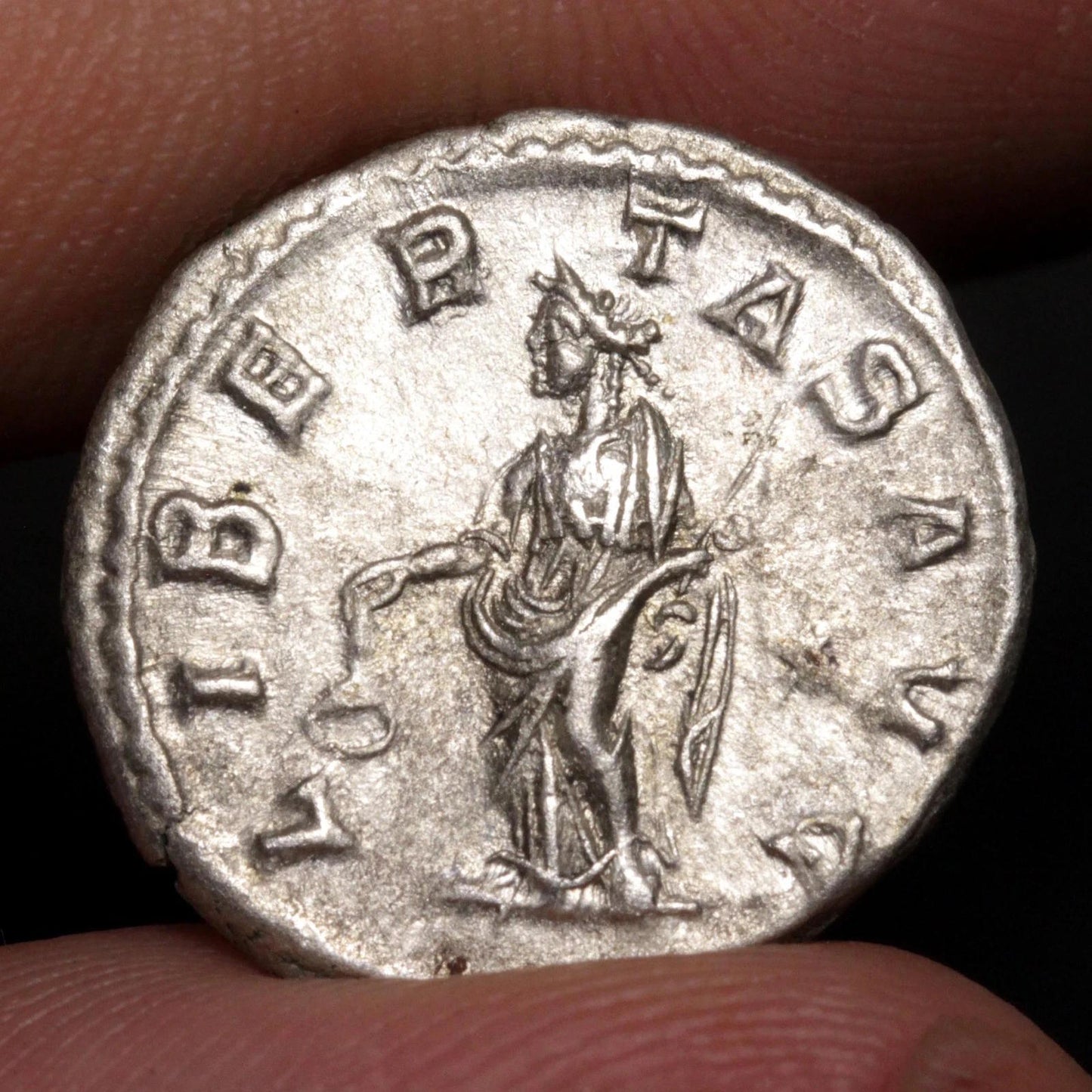 An ancient silver coin held between fingers, showing a relief of a standing figure surrounded by the inscription 'LIBER TAS AVG'.