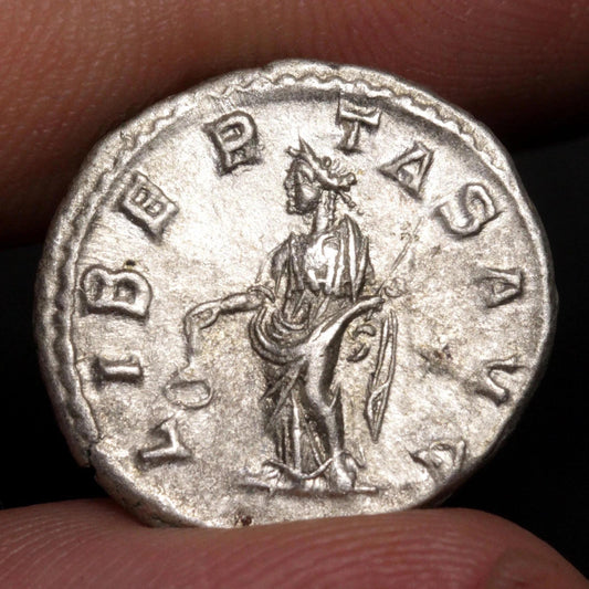An ancient silver coin held between fingers, showing a relief of a standing figure surrounded by the inscription 'LIBER TAS AVG'.