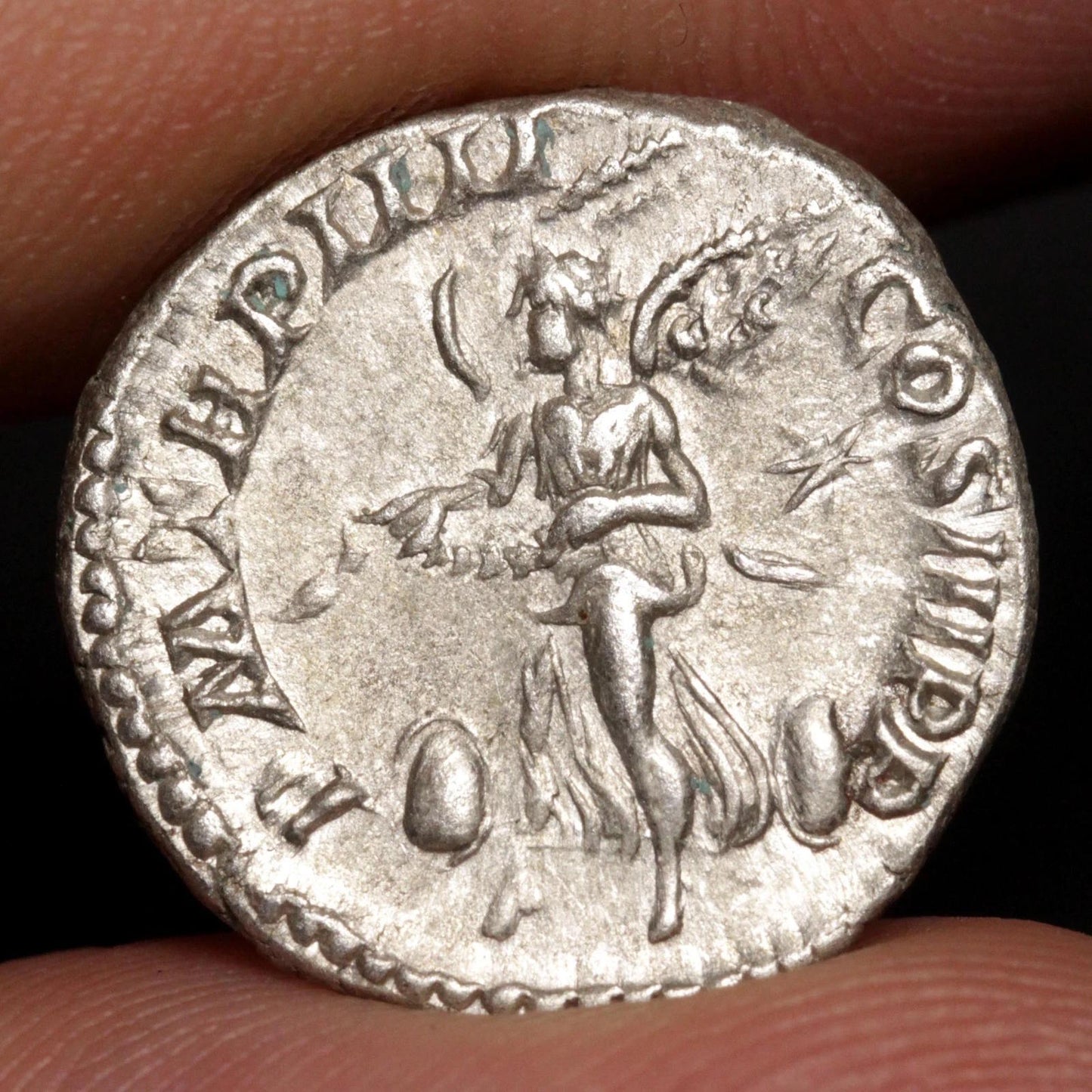 An ancient silver coin held between fingers, showing a relief of a winged female figure with inscriptions around the edge.