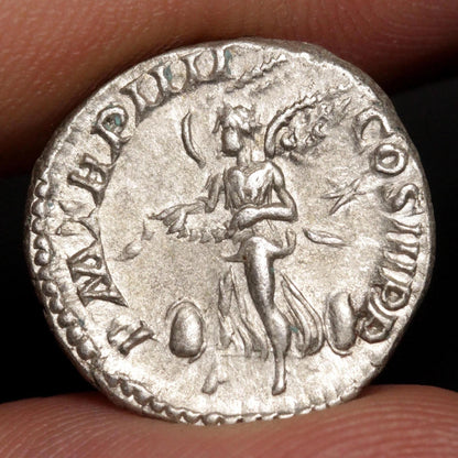 An ancient silver coin held between fingers, showing a relief of a winged female figure with inscriptions around the edge.