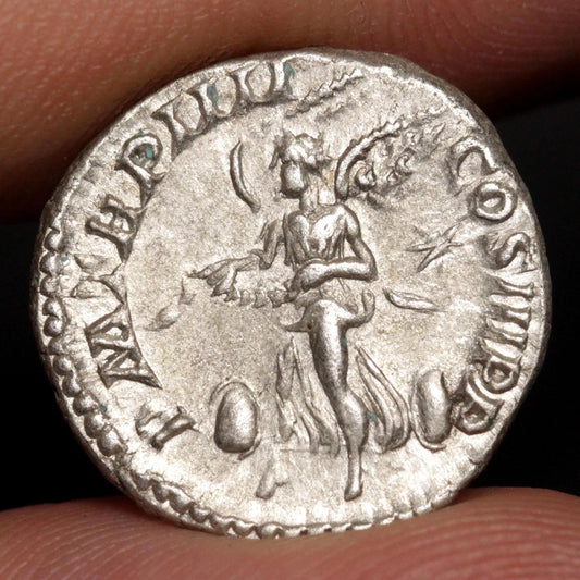 An ancient silver coin held between fingers, showing a relief of a winged female figure with inscriptions around the edge.
