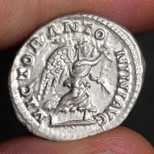 An ancient silver coin held between two fingers, showing a detailed relief of a winged figure and raised Roman text around the edge.