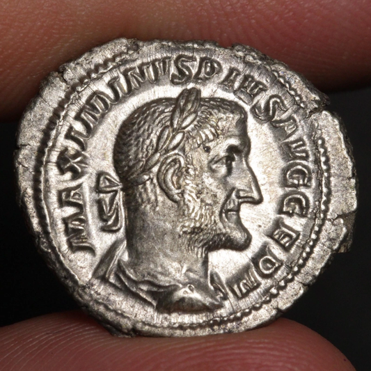 An ancient silver Roman coin featuring a profile portrait of a bearded man with a laurel wreath, held between two fingers against a dark background.