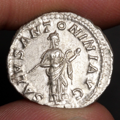 An ancient silver coin held between fingers, featuring a standing figure and raised Latin text around the edge.